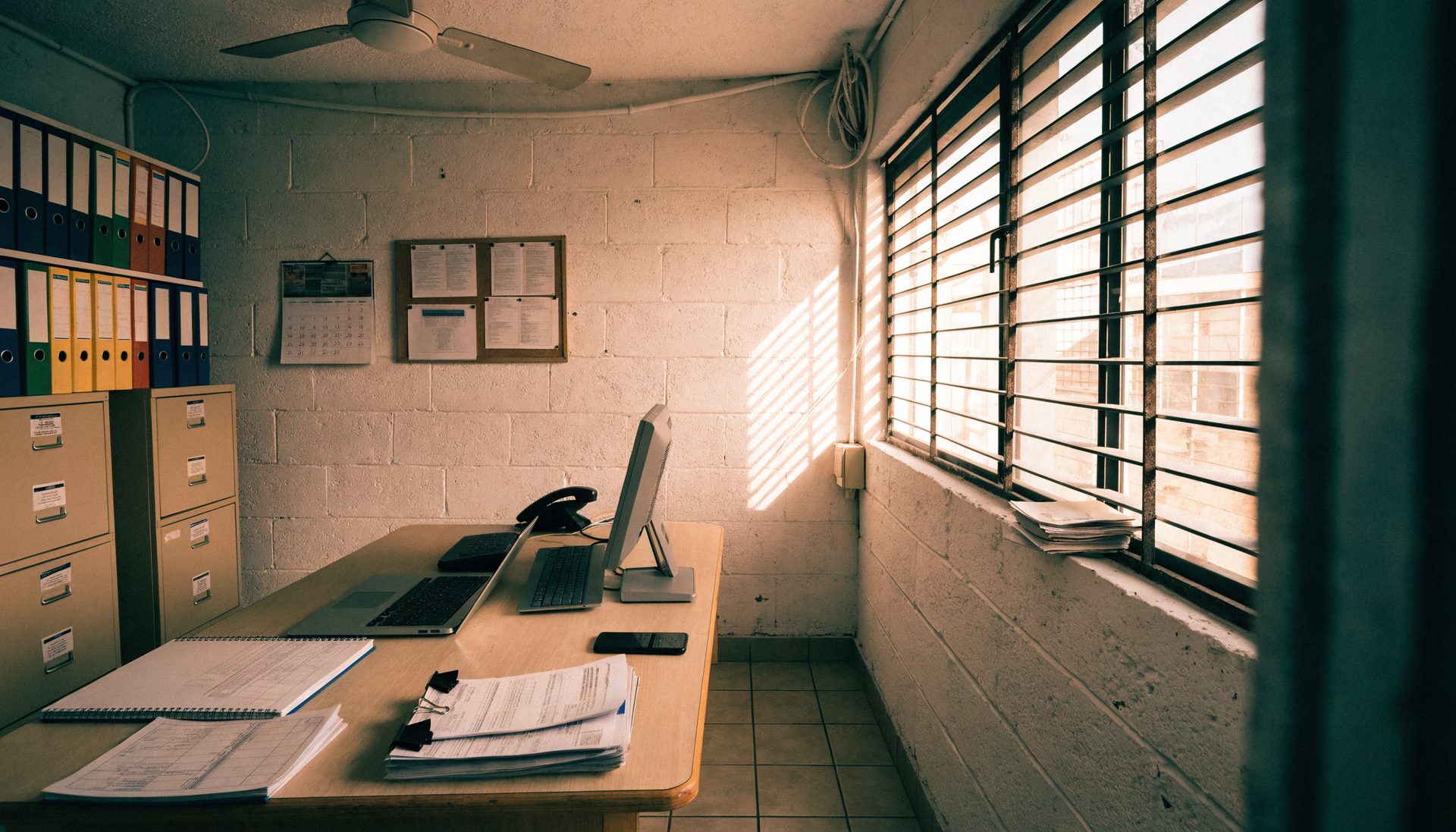 A working back office with a laptop, an older monitor, a ledger book, stacks of printed forms and metal filing cabinets, lit through a louvred window.