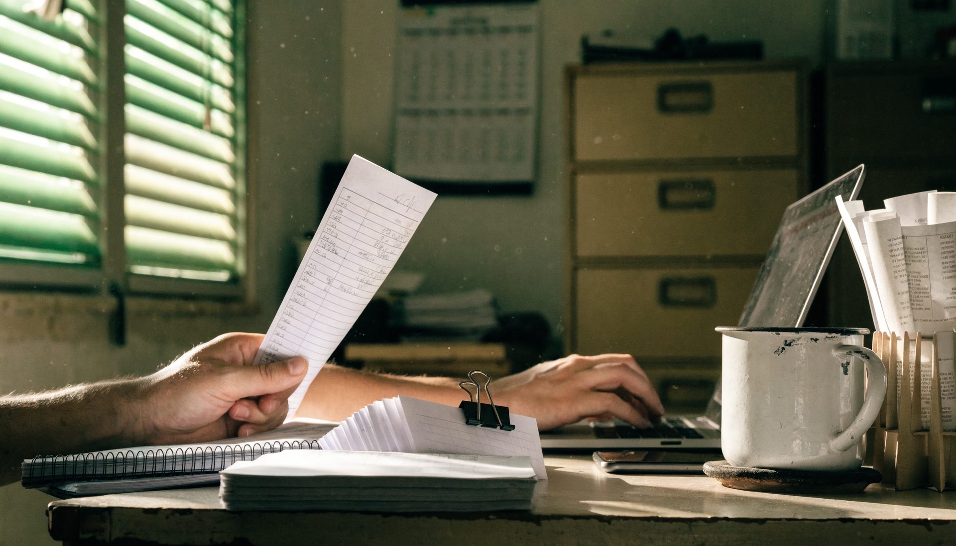 Hands copying details from a printed paper docket into a laptop at a cluttered office desk.