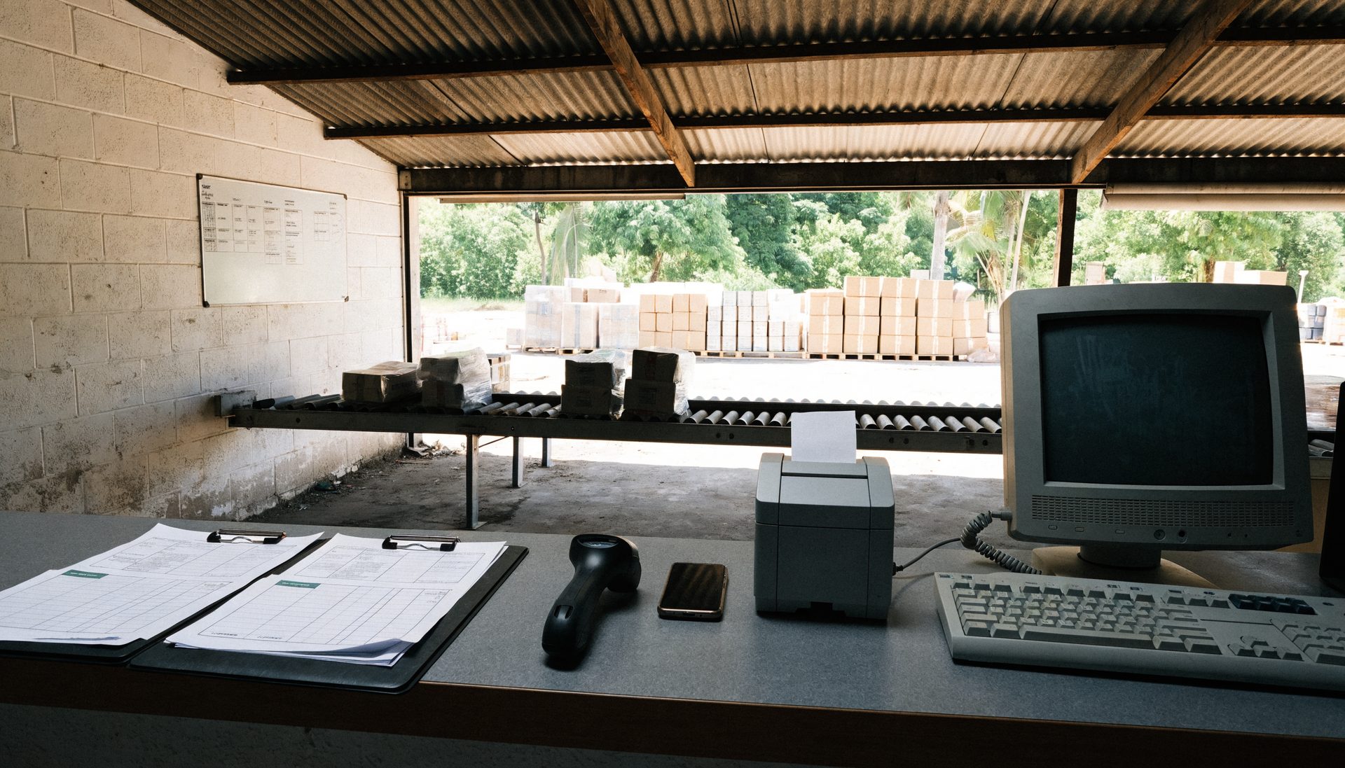 A depot service counter where carbon-copy dockets, a barcode scanner, a smartphone and an older desktop monitor are used side by side.